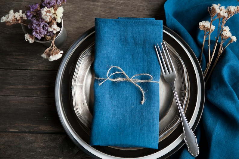 May include: A blue linen napkin tied with twine, placed on a silver and black plate with a fork. The plate is on a wooden table with a blue linen tablecloth and dried flowers.