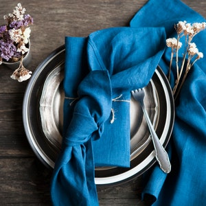Peut inclure: Une serviette en lin bleu est pli&eacute;e et attach&eacute;e avec de la ficelle sur une assiette argent&eacute;e et noire. Une fourchette est plac&eacute;e sur l'assiette. La serviette est entour&eacute;e de fleurs s&eacute;ch&eacute;es.