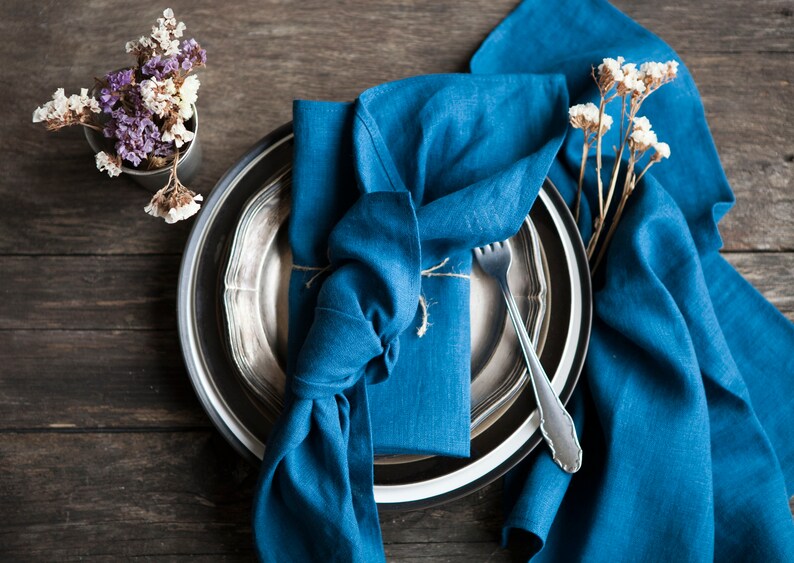 Peut inclure: Une serviette en lin bleu est pli&eacute;e et attach&eacute;e avec de la ficelle sur une assiette argent&eacute;e et noire. Une fourchette est plac&eacute;e sur l'assiette. La serviette est entour&eacute;e de fleurs s&eacute;ch&eacute;es.