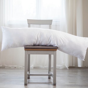 May include: A white body pillow resting on a gray chair with a wooden seat. The chair is in a room with a window and white curtains.