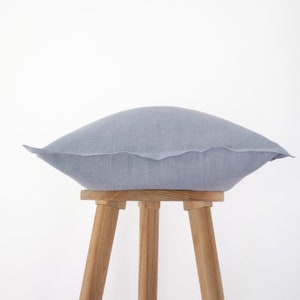 Peut inclure: Un coussin en lin gris clair pos&eacute; sur un tabouret en bois &agrave; trois pieds. Le coussin est carr&eacute; et de design simple.