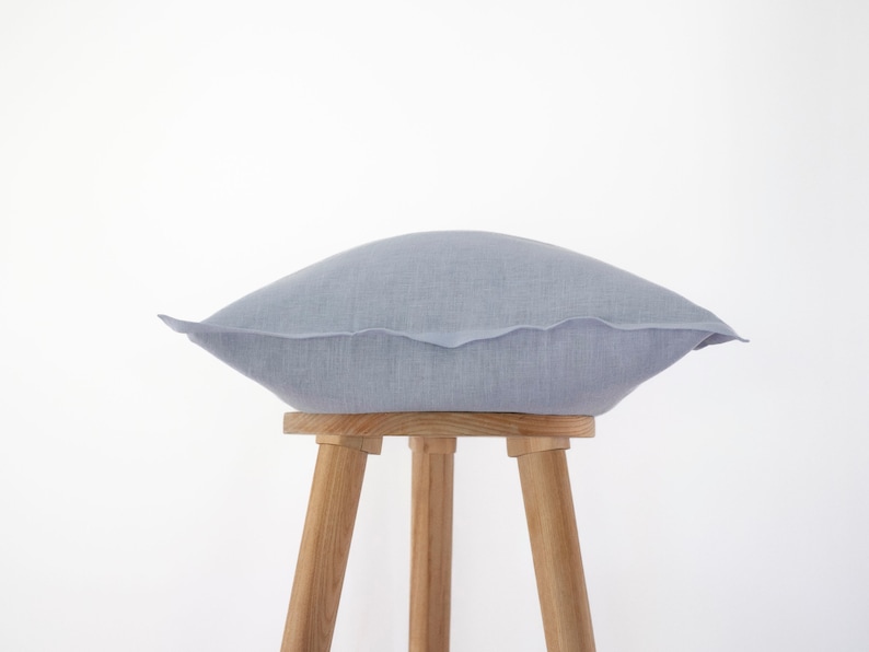 Peut inclure: Un coussin en lin gris clair pos&eacute; sur un tabouret en bois &agrave; trois pieds. Le coussin est carr&eacute; et de design simple.