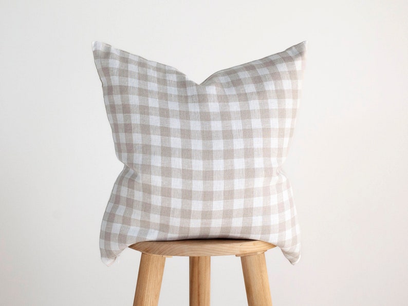May include: A beige and white gingham pillow sits on a wooden stool. The pillow is made of a soft, textured fabric and has a square pattern.