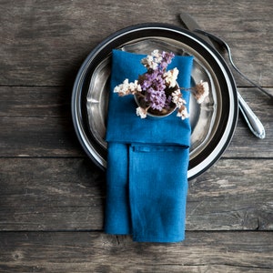 Peut inclure: Une serviette en lin bleu est pli&eacute;e et plac&eacute;e sur une assiette de d&icirc;ner argent&eacute;e et noire. Un petit bouquet de fleurs violettes et blanches est plac&eacute; sur la serviette.