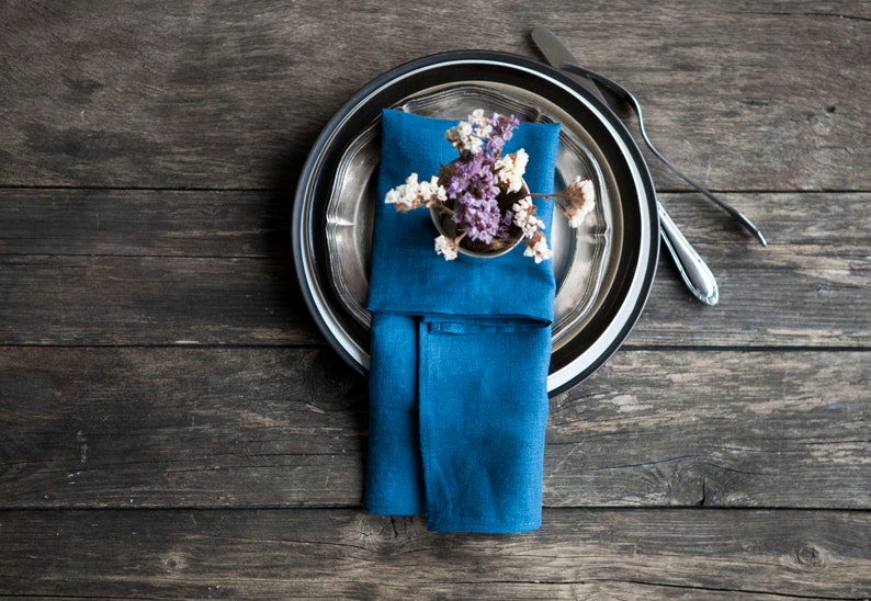 Peut inclure: Une serviette en lin bleu est pli&eacute;e et plac&eacute;e sur une assiette de d&icirc;ner argent&eacute;e et noire. Un petit bouquet de fleurs violettes et blanches est plac&eacute; sur la serviette.