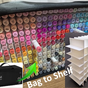May include: A black carrying case filled with numerous colourful alcohol markers, each featuring a cap with a specific colour code and name. A white grid-style plastic organiser insert is shown separately, designed to hold markers upright in a shelf format.