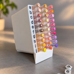 May include: A white plastic marker organiser stand holding multiple coloured markers. The side of the stand features a vertical column of white tabs labelled with the lowercase letters a through j in black text. Several loose white square caps are nearby.