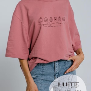 Può includere: Una t-shirt rosa polvere con una fila di piccole icone a linee nere che includono una borsa della spesa, cuffie, flacone di profumo, tazza di caffè, candela e cupcake, con il testo "quality me time" e "SHOPPING GOOD MUSIC SLOW MOMENTS".