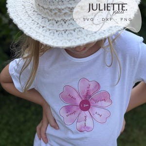 May include: A white t-shirt featuring a pink watercolor flower graphic. The center of the flower reads 'I am' and the petals contain the words 'brave', 'beautiful', 'strong', 'kind', 'unique', and 'enough'. A white lace hat is worn above the shirt.