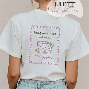 Pode incluir: Uma t-shirt branca com uma borda retangular festonada roxa contendo um gráfico de uma chávena de café com pequenos corações roxos. O texto diz: bring me coffee and tell me I'm pretty. O design está centrado nas costas da t-shirt.