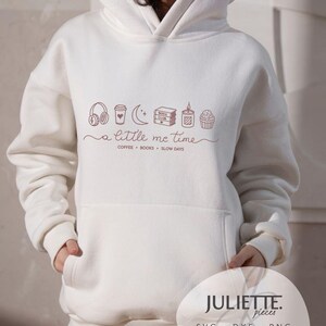 Puede incluir: Una sudadera con capucha blanca con un estampado gráfico granate de auriculares, una taza de café, una luna creciente, libros, una vela y un cupcake. El texto dice a little me time, COFFEE, BOOKS, SLOW DAYS.