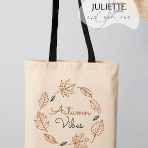 Può includere: Una borsa tote in tela beige con spallacci neri caratterizzata da un design a corona circolare composto da illustrazioni di foglie autunnali arancioni e nere. Il testo Autumn Vibes è stampato al centro con caratteri arancioni e neri.