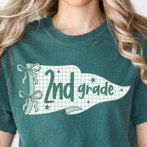 May include: A dark green short-sleeve t-shirt featuring a white pennant graphic with a light green gingham pattern. The graphic includes the text 2nd grade in dark green script, accented by two bows on the left side and small star shapes.