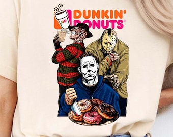 May include: A cream-colored short-sleeve t-shirt featuring a graphic print of three horror movie characters eating donuts. The text Dunkin' Donuts is printed above the characters, who are holding a coffee cup and a plate of assorted donuts.