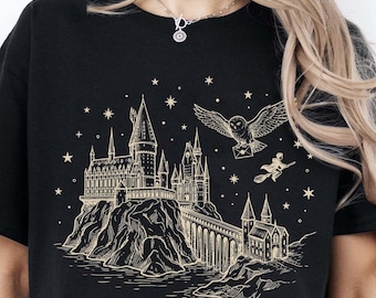 May include: A black short-sleeved t-shirt featuring a gold-toned graphic print of a large castle perched on a rocky cliff, a bridge, a flying owl carrying a letter, a figure on a broomstick, and scattered stars against a dark background.