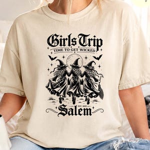May include: An ivory short-sleeve t-shirt featuring a black graphic print of three witches holding hands under a full moon. The text reads Girls Trip, Time to Get Wicked, and Salem, Est. 1692. The design includes stars, bats, and a broomstick.