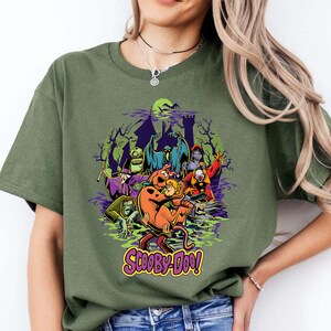 May include: An olive green t-shirt featuring a graphic print of Scooby-Doo and Shaggy surrounded by various villains from the series, including a ghost, a knight, and a clown, set against a dark castle silhouette with the text Scooby-Doo below.
