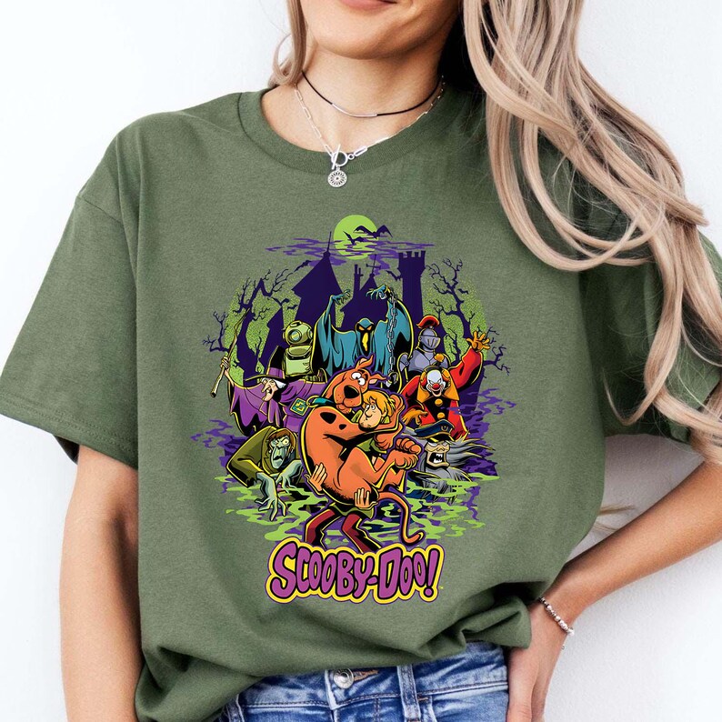 May include: An olive green t-shirt featuring a graphic print of Scooby-Doo and Shaggy surrounded by various villains from the series, including a ghost, a knight, and a clown, set against a dark castle silhouette with the text Scooby-Doo below.