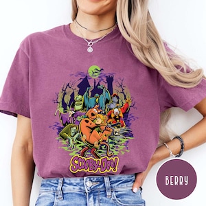 May include: A berry-colored short-sleeve t-shirt featuring a graphic print of Scooby-Doo and Shaggy surrounded by various ghosts and monsters in front of a haunted castle silhouette. The text Scooby-Doo is printed in yellow at the bottom.