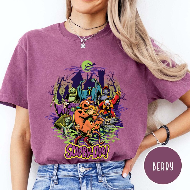 May include: A berry-colored short-sleeve t-shirt featuring a graphic print of Scooby-Doo and Shaggy surrounded by various ghosts and monsters in front of a haunted castle silhouette. The text Scooby-Doo is printed in yellow at the bottom.