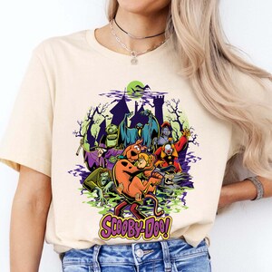 May include: A cream-colored short-sleeve t-shirt featuring a colorful graphic of Scooby-Doo and Shaggy surrounded by various classic villains from the series. The design includes a haunted castle silhouette, green mist, and the Scooby-Doo logo in purple.