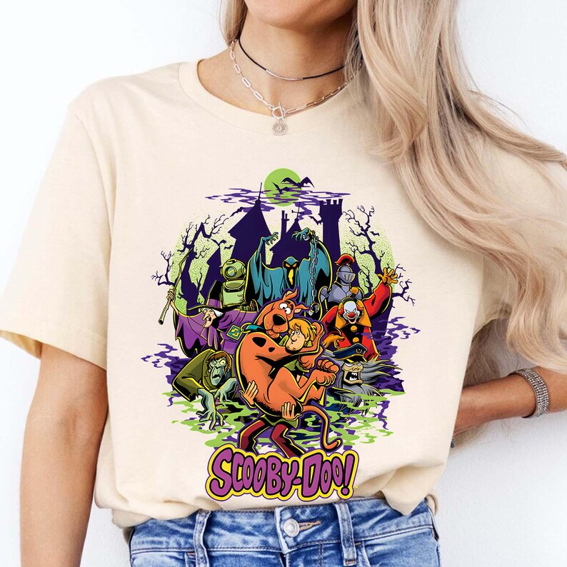 May include: A cream-colored short-sleeve t-shirt featuring a colorful graphic of Scooby-Doo and Shaggy surrounded by various classic villains from the series. The design includes a haunted castle silhouette, green mist, and the Scooby-Doo logo in purple.