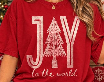 May include: A red short-sleeved t-shirt featuring a large white graphic print. The design displays the word JAY with a stylized evergreen tree replacing the letter A, and the text to the world printed in a smaller script font below.