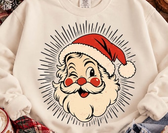 May include: A cream-colored crewneck sweatshirt featuring a graphic print of a winking Santa Claus face with a red hat, white beard, and rosy cheeks, surrounded by black radiating lines. The inner collar tag displays the Comfort Colors brand name.