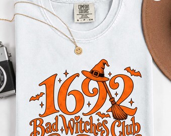 May include: A white t-shirt featuring an orange graphic with the text 1692 Bad Witches Club. The design includes a witch hat, a broomstick, small bats, and star accents. The shirt has a Comfort Colors label at the neck and a crew neckline.