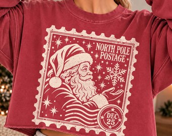 May include: A dark red long-sleeve cropped sweatshirt featuring a cream-colored graphic of a postage stamp. The design includes Santa Claus, a snowflake, stars, and the text North Pole Postage and Dec 25 with a distressed texture.
