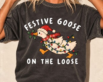 May include: A charcoal gray t-shirt featuring a graphic of a white goose wearing a red Santa hat and scarf, holding a sprig of holly, and tangled in colorful Christmas lights. The text reads FESTIVE GOOSE ON THE LOOSE in white letters.
