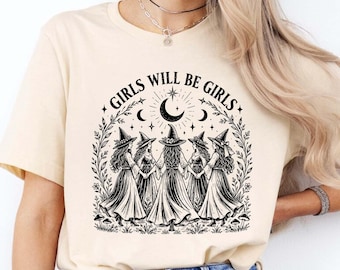 May include: A cream-colored short-sleeve t-shirt featuring a black graphic print of five figures wearing witch hats and long dresses standing in a circle. The text GIRLS WILL BE GIRLS is arched above the figures, with a crescent moon and stars.