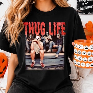 May include: A black t-shirt featuring the red text THUG LIFE above a graphic of four horror movie characters sitting on porch steps. The characters include figures wearing masks resembling Michael Myers, Ghostface, and Jason Voorhees.
