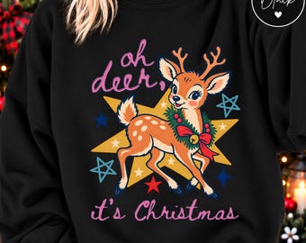 May include: A black sweatshirt featuring a graphic of a fawn wearing a green wreath with a red bow. Behind the fawn is a large yellow star and several smaller blue and red stars. The text reads "oh deer, it's Christmas" in pink lettering.