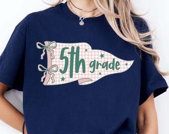 May include: A navy blue t-shirt featuring a graphic of a pennant flag with a pink and white gingham pattern. The flag displays the text 5th grade in green, accented with two pink bows on the left side and small green stars scattered across the design.