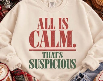 May include: A cream-colored crewneck sweatshirt featuring the red text ALL IS CALM. followed by two horizontal lines and the green text THAT'S SUSPICIOUS. The garment has long sleeves, a ribbed collar, and a Comfort Colors brand label at the neck.
