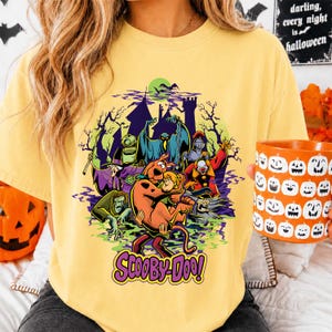 May include: A pale yellow t-shirt featuring a graphic print of Scooby-Doo and Shaggy surrounded by various monsters, a haunted castle, and trees. The text 'SCOOBY-DOO!' is printed in purple at the bottom of the design. The shirt has a crew neckline.