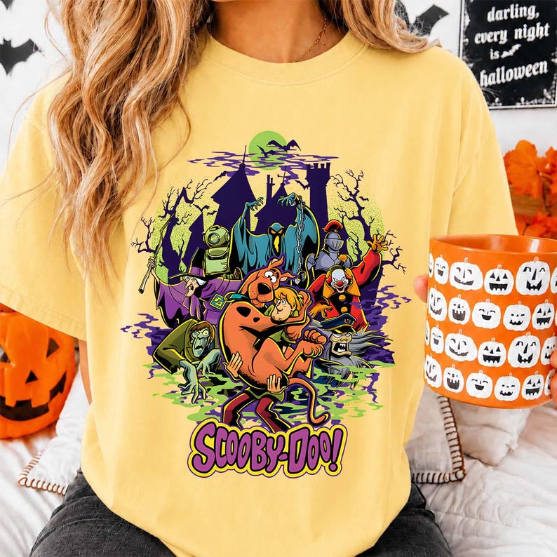May include: A pale yellow t-shirt featuring a graphic print of Scooby-Doo and Shaggy surrounded by various monsters, a haunted castle, and trees. The text 'SCOOBY-DOO!' is printed in purple at the bottom of the design. The shirt has a crew neckline.