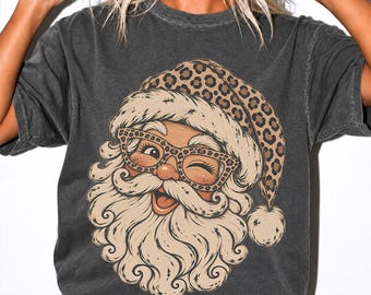 May include: A charcoal gray short-sleeve t-shirt featuring a graphic print of Santa Claus wearing a leopard print hat and matching leopard print glasses. The illustration shows Santa with a large white beard, winking, and smiling.