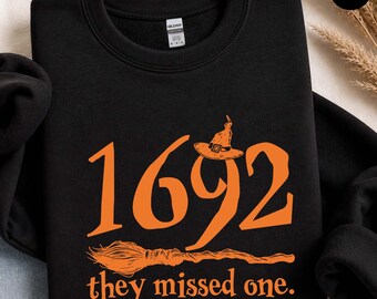 May include: A black crewneck sweatshirt featuring orange graphic text that reads 1692 they missed one. The design includes an orange witch hat perched atop the number nine and an orange broomstick illustration positioned below the text.