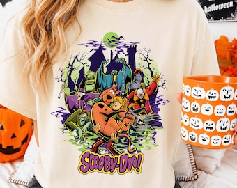 May include: A cream-colored short-sleeve t-shirt featuring a Scooby-Doo graphic print. The design displays Scooby-Doo and Shaggy surrounded by various ghosts and monsters in front of a haunted castle under a green moon, with the text Scooby-Doo below.