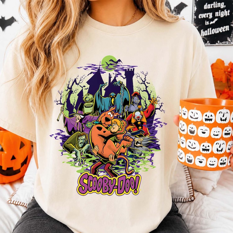 May include: A cream-colored short-sleeve t-shirt featuring a Scooby-Doo graphic print. The design displays Scooby-Doo and Shaggy surrounded by various ghosts and monsters in front of a haunted castle under a green moon, with the text Scooby-Doo below.