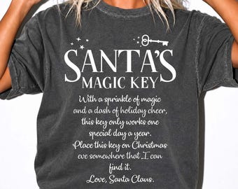 May include: A charcoal gray short-sleeve t-shirt featuring white text that reads SANTA'S MAGIC KEY followed by a poem about Christmas Eve and a graphic of a key. The shirt has a crew neckline and a relaxed fit with a washed-out fabric appearance.