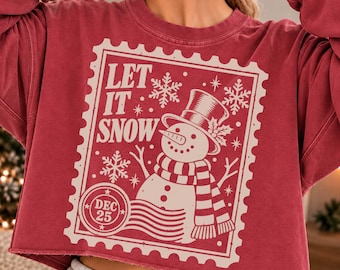 May include: A dark red cropped sweatshirt featuring a cream-colored graphic of a postage stamp. The design includes a snowman wearing a top hat and scarf, snowflakes, and the text "LET IT SNOW" with a "DEC 25" postmark circle.