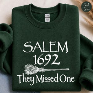 Peut inclure: Un sweat-shirt à col rond vert forêt avec un texte imprimé en blanc indiquant SALEM 1692 They Missed One, accompagné d'une illustration graphique d'un balai placée entre les lignes de texte sur le devant du vêtement.