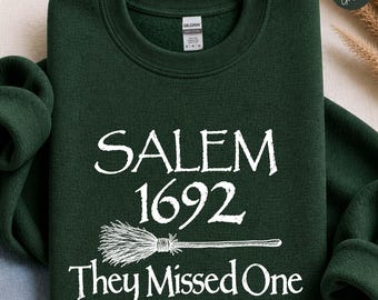 May include: A forest green crewneck sweatshirt featuring white screen-printed text that reads SALEM 1692 They Missed One, accompanied by a graphic illustration of a broomstick centered between the lines of text on the front of the garment.
