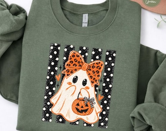 May include: A military green crewneck sweatshirt featuring a graphic of a white ghost wearing a large orange leopard print bow. The ghost holds a small orange jack-o'-lantern bucket. The design is set against black vertical stripes with white polka dots.