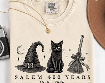 May include: An ivory t-shirt featuring a black ink graphic of a witch hat, a black cat, and a broomstick. Below the illustrations, the text reads SALEM 400 YEARS 1626 - 2026. The shirt has a Comfort Colors label at the neck and a crew neckline.