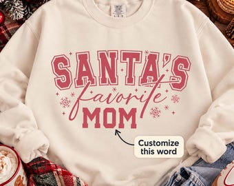 May include: A cream-colored crewneck sweatshirt featuring the red text SANTA'S favorite MOM in a mix of block and script fonts, accented with small star and snowflake graphics. A small white box with the text Customize this word is included.