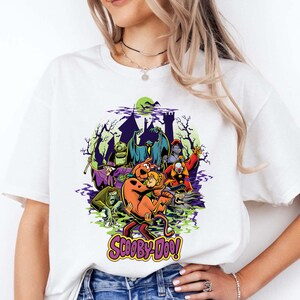 May include: A white t-shirt featuring a colorful graphic print of Scooby-Doo characters and various monsters in front of a dark castle silhouette. The design includes a green moon, purple smoke, and the text Scooby-Doo in a stylized yellow font.